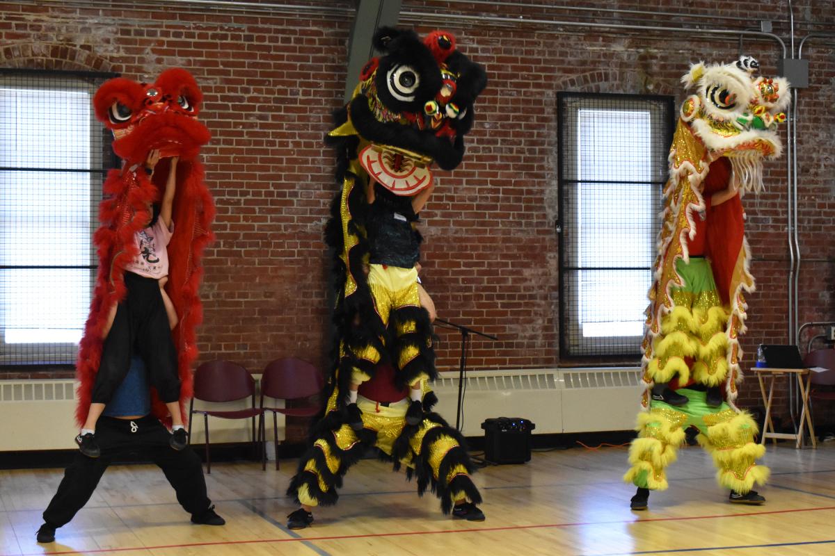 three children wear tall Lion Dance costumes in red, black, yellow, and gold